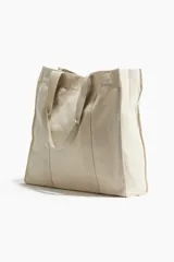Bolso shopper de lona color beige con doble asa de hombro y cierre de botón a presión. Tiene un compartimento exterior y un compartimento interior pequeño, ambos abiertos.