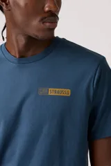 Remera de manga corta, cuello redondo, color azul oscuro, con un pequeño parche gráfico rectangular con texto en tono mostaza en el pecho.