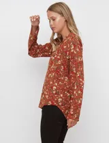 Blusa de manga larga color marrón terracota con estampado floral en tonos rojos, blancos y verdes. Presenta un escote en V con cuello mao y mangas que se pueden ajustar con presilla y botón.