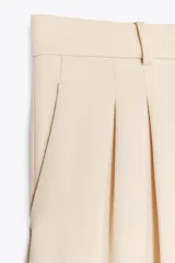 Bermuda color crema de tiro medio con trabillas, bolsillos laterales, pinzas delanteras y cierre frontal con cremallera, botón y gancho metálico.