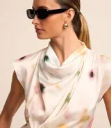 Blusa sin mangas de tela satinada color blanco, con cuello drapeado, lazo lateral y estampado de manchas en tonos rosados, amarillos y verdes.