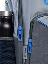 Mochila deportiva Umbro modelo Zeeva, color gris con detalles en azul eléctrico. Presenta el logo Umbro grande en azul en el panel frontal superior y un panel inferior texturizado con patrón de logo repetido. Incluye bolsillo lateral de malla para botella y tiradores de cierre en azul.