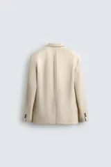 Blazer color beige confeccionado en tejido con lana, de corte relajado. Tiene cuello con solapas de muesca, manga larga con puños abotonados, bolsillo de vivo en el pecho, bolsillos de solapa en la cadera, bolsillo interior, bajo con abertura en el centro de la espalda y cierre frontal con botones.