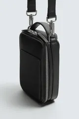 Bandolera negra con textura grabada, forma rectangular y cierre de cremallera. Tiene un bolsillo interior plano y otro frontal plano. Incluye asa de hombro ajustable y extraíble.