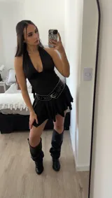 Minivestido negro con escote halter, espalda descubierta, falda con volados asimétricos, cinturón con tachas y botas de caña alta.
