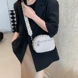 Cartera bandolera pequeña color beige, de cuero sintético con textura granulada. Tiene correa ancha de tela estampada con motivos geométricos y cierre superior con cremallera. Incluye un bolsillo frontal con cremallera y un dije metálico.