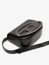 Bandolera mediana de cuero liso color blanco, con solapa frontal que presenta una placa de metal esmaltado con el logo D ovalado en relieve. Incluye correa ajustable para llevar al hombro o cruzada, y compartimento interior amplio con dos secciones planas adicionales.