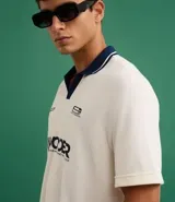 Remera de manga corta con cuello polo contrastante en azul marino y abertura en V. El cuerpo de la prenda es color crema y lleva un estampado frontal con la palabra "WONDER".