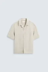 Camisa color beige claro de lino, corte relaxed fit, cuello bowling, manga corta, bajo con aberturas laterales y cierre frontal con botones.
