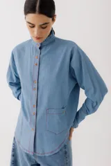 Camisa celeste de jean con cuello alto con volados y mangas largas abullonadas con puños con volados. Presenta botones de madera y costuras rojas a contraste.