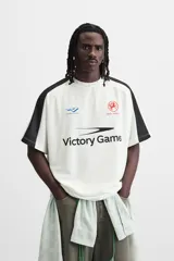 Camiseta blanca de corte holgado con mangas cortas negras y cuello redondo. Presenta estampados frontales con logos y textos en azul y rojo.