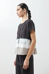 Blusa de manga corta con cuello redondo y cierre frontal parcial con botones. Presenta un diseño tie dye en tonos grises y blancos en la parte inferior.