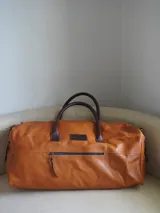 Bolso de viaje tipo weekender de cuero color marrón con doble asa de mano y cierre superior con cremallera. Tiene un bolsillo externo con cremallera y bolsillos internos.