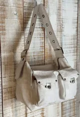 Bolso bandolera color beige con textura símil cuero de cocodrilo. Tiene correa de hombro ajustable con ojales metálicos, cierre superior con cremallera y dos bolsillos frontales con cierre de broche metálico.