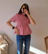 Blusa de cuello alto con diseño de cuadros vichy en rojo y blanco. Presenta un cuerpo elástico con nido de abeja (smocking), mangas cortas y un bajo con volado tipo peplum.