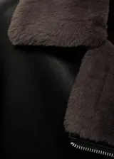 Chaqueta negra de cuero sintético con cuello de piel sintética gris, cierre cruzado con cremallera y bolsillos laterales con cremallera.