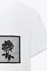 Camiseta blanca de algodón con cuello redondo y mangas cortas. Presenta un bolsillo de tul con una flor bordada y abalorios en el delantero.