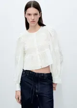 Blusa blanca con bordado floral, cuello redondo, mangas largas abullonadas con puños abotonados, cierre frontal con botones y corte crop.