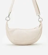 Bolso mediano color crema, con forma de media luna y correa ajustable con aros metálicos.