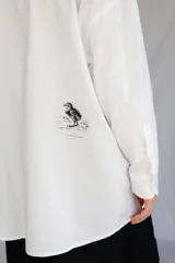 Camisa blanca de algodón con cuello con lengüeta y botón labrado, manga larga con puños y botones, corte oversize y estampado de aves en el delantero.