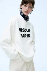 Buzo blanco de corte holgado con cuello redondo y puños y ruedo acanalados. Estampa frontal con la frase "Bisous Paris" en letras negras.