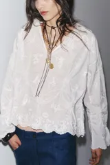 Blusa blanca de algodón con cuello solapa, escote pico y manga larga acampanada. Detalle de frunces y bordados a tono.