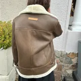 Campera gamulán color marrón con interior de piel sintética color crema, cierre frontal con cremallera descentrada, cuello con solapas amplias y bolsillos laterales.