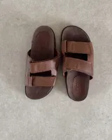 Sandalias bajas con base marrón y dos tiras anchas de cuero sintético negro con ajuste de velcro.