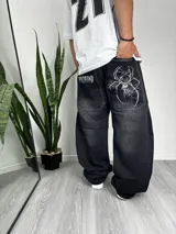Pantalón baggy negro de denim con bolsillos traseros con bordado de calavera con telaraña y logo de la marca en blanco.