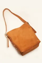 Cartera tipo balde de gamuza color marrón, con correa ajustable decorada con tachas metálicas plateadas y logo grabado en la parte frontal.
