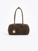 Cartera rectangular de estructura firme confeccionada en cuero descarne vacuno color marrón. Presenta manijas fijas, cierre superior y un charm metálico dorado en forma de vaquita de San Antonio colgando de un tag lateral. Incluye el logo de la marca grabado en relieve en el frente.