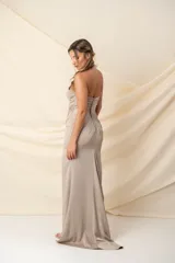 Vestido largo de fiesta color arena, de crepé, con escote asimétrico y un solo hombro. Presenta una moña con aplique bordado en el hombro y la cintura. Más largo atrás.