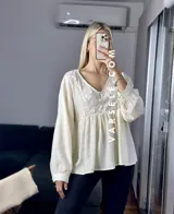 Blusa color crema con escote en V, mangas largas abullonadas con puños elásticos y detalles de encaje y bordado en el frente.