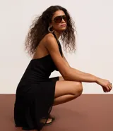 Vestido corto negro de tejido liviano, con escote halter que forma un drapeado (escote degagê) en el busto y ruedo asimétrico con volados.