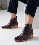 Botín de cuero color marrón chocolate, con puntera fina y cierre lateral. Presenta un detalle de cuero con textura de cocodrilo en el empeine. El taco es de madera con folia de cuero y la suela es de caucho. La caña mide 12cm.