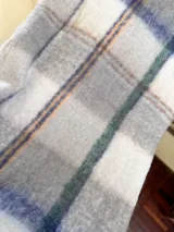 Bufanda rectangular con estampado a cuadros en tonos gris, blanco, azul y verde. Tiene flecos en los extremos.