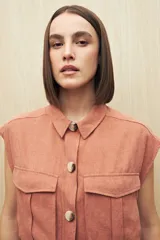 Chaleco color beige con cuello camisero, sin mangas, cierre frontal con botones y cinturón de lazo. Presenta bolsillos de parche en el pecho y pinzas verticales en el frente.
