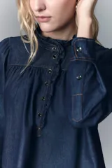 Camisa de denim azul oscuro con cuello mao, corte recto, mangas largas con puños abotonados, cierre frontal con botones a presión y detalle de volados en el cuello y la pechera.