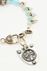Pulsera de cuentas de piedras semipreciosas en tonos rojizos, negros y verde claro, con cierre de mosquetón dorado y un dije colgante en forma de escudo plateado con una cruz en relieve.