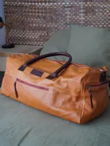 Bolso de viaje tipo weekender de cuero color marrón con herrajes metálicos y forro interior. Cuenta con doble asa de mano, correa de hombro ajustable y extraíble, cierre superior con cremallera, bolsillos internos y externos.