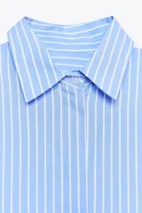 Camisa celeste a rayas verticales blancas, de cuello solapa y manga tres cuartos. Tiene aberturas laterales en el ruedo y cierre frontal con botones ocultos.
