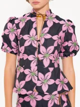 Top con estampado de flores rosas y verdes sobre fondo azul marino, cuello mao con jareta y tiras de gamuza para anudar. Escote en forma de gota, acceso mediante botones forrados en tela base, corte en cintura que le da vuelo a la cadera y mangas abullonadas con elástico en el puño.