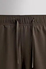 Pantalón de jogging color marrón oscuro, corte regular fit, confeccionado en tejido técnico ligero y elástico. Cuenta con cintura elástica ajustable con cordón, bolsillos frontales con cierre de cremallera, bolsillo trasero con cierre de cremallera oculta en costura y bajo ajustable con elástico.