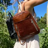 Mochila de cuero color marrón con textura tipo cocodrilo en el bolsillo frontal. Tiene cuatro compartimentos independientes con cierre, dos bolsillos internos y correas de cuero regulables.