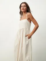 Vestido largo color beige claro con escote cuadrado y tirantes finos anudados en los hombros. Presenta un corte imperio con frunce en la cintura y falda amplia con bolsillos laterales.