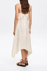 Vestido midi de algodón y lino color crudo, corte línea A con vuelo y bajo irregular. Presenta escote recto con tirantes finos y top superpuesto con bordados a contraste.