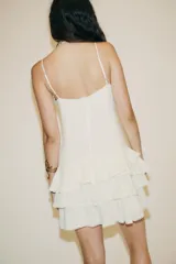 Vestido corto color crudo, de gasa, con escote en V y tirantes finos regulables. Presenta cordón cruzado con lazada en el delantero y falda con volantes. Cierre trasero con cremallera oculta.