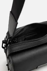 Bandolera negra con textura grabada, compartimento principal con cierre de cremallera, bolsillo interior con cremallera, bolsillo frontal con solapa y cremallera, asa de hombro removible y ajustable.