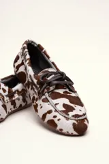 Mocasín tipo boat shoe con estampado de vaca en tonos marrón y blanco, confeccionado en cuero. Cuenta con cordones de cuero marrón oscuro y suela de goma.