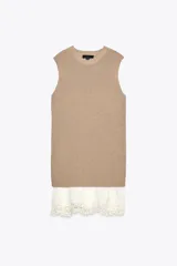 Vestido color beige, de corte recto, con cuello redondo y sin mangas. Presenta un tejido de punto en la parte superior y un volante de encaje blanco en el ruedo.